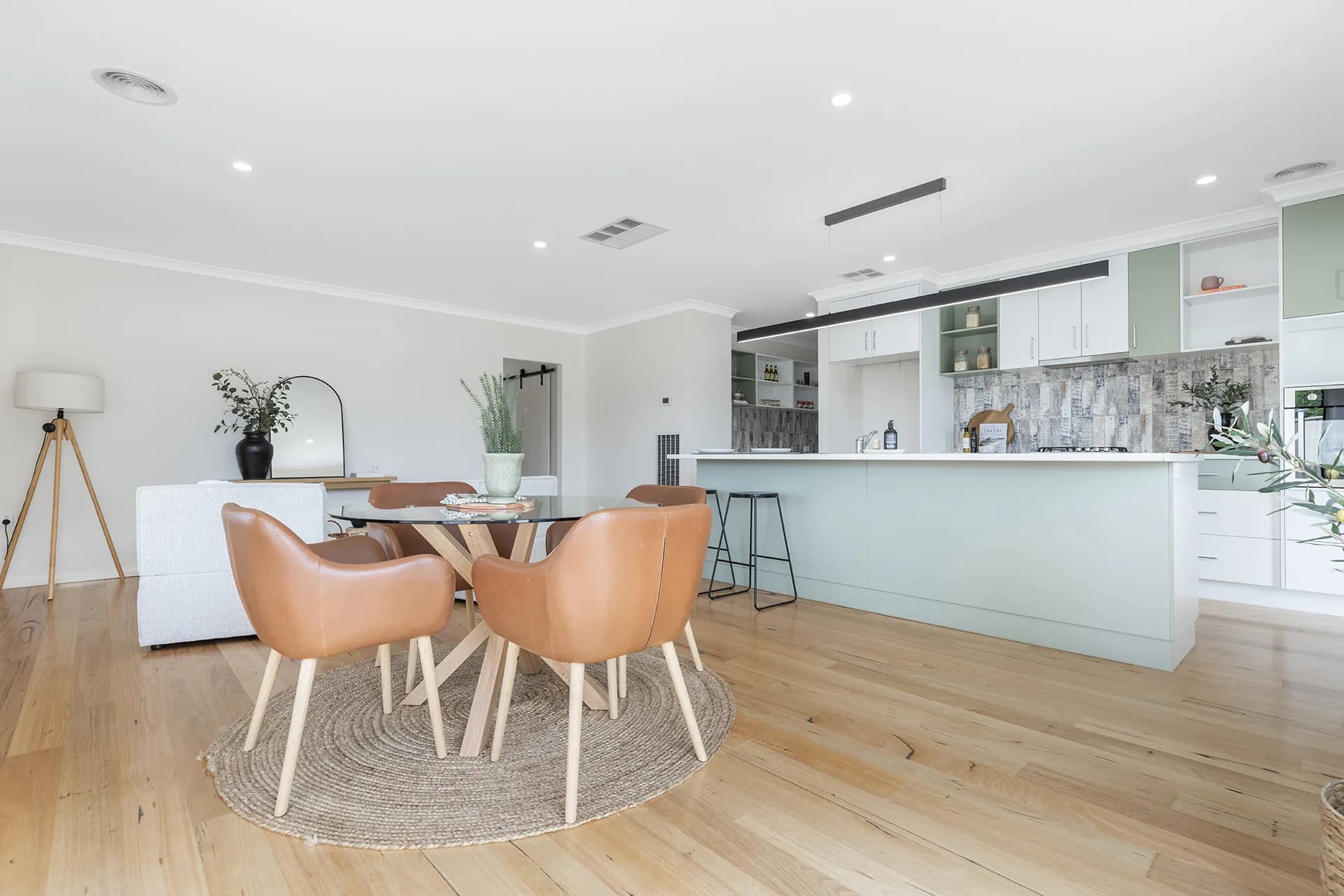 Dining table with tan leather chairs on a jute rug beside a sage green kitchen island at Maker Parade, Echuca